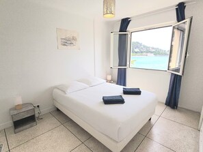 Ein Hotelzimmer mit einem Bett, einem Nachttisch, einer Lampe, einem Fenster mit Blick und einem an der Wand hängenden Kunstwerk.