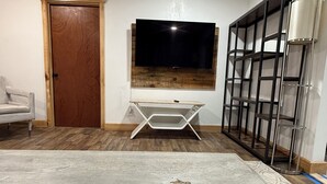 Une pièce avec une porte en bois, une chaise pliante blanche, un téléviseur fixé au mur en bois et une étagère en métal.