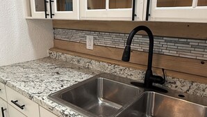 Une cuisine avec des armoires blanches, un évier en acier inoxydable à deux bacs, des comptoirs en granit et un dosseret avec un motif en briques.