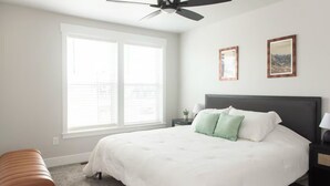 Quarto com cama, mesa de cabeceira, banquinho, ventoinha no teto e duas quadros na parede.