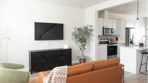 Sala de estar com sofá, televisão, planta em vaso e área de cozinha.