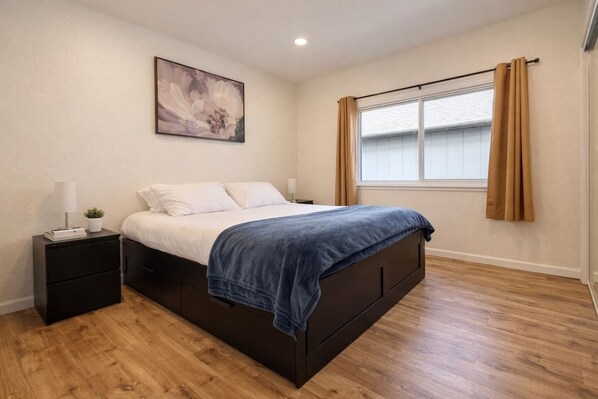 A bedroom with a bed, a nightstand, a window with curtains, and a painting on the wall.
