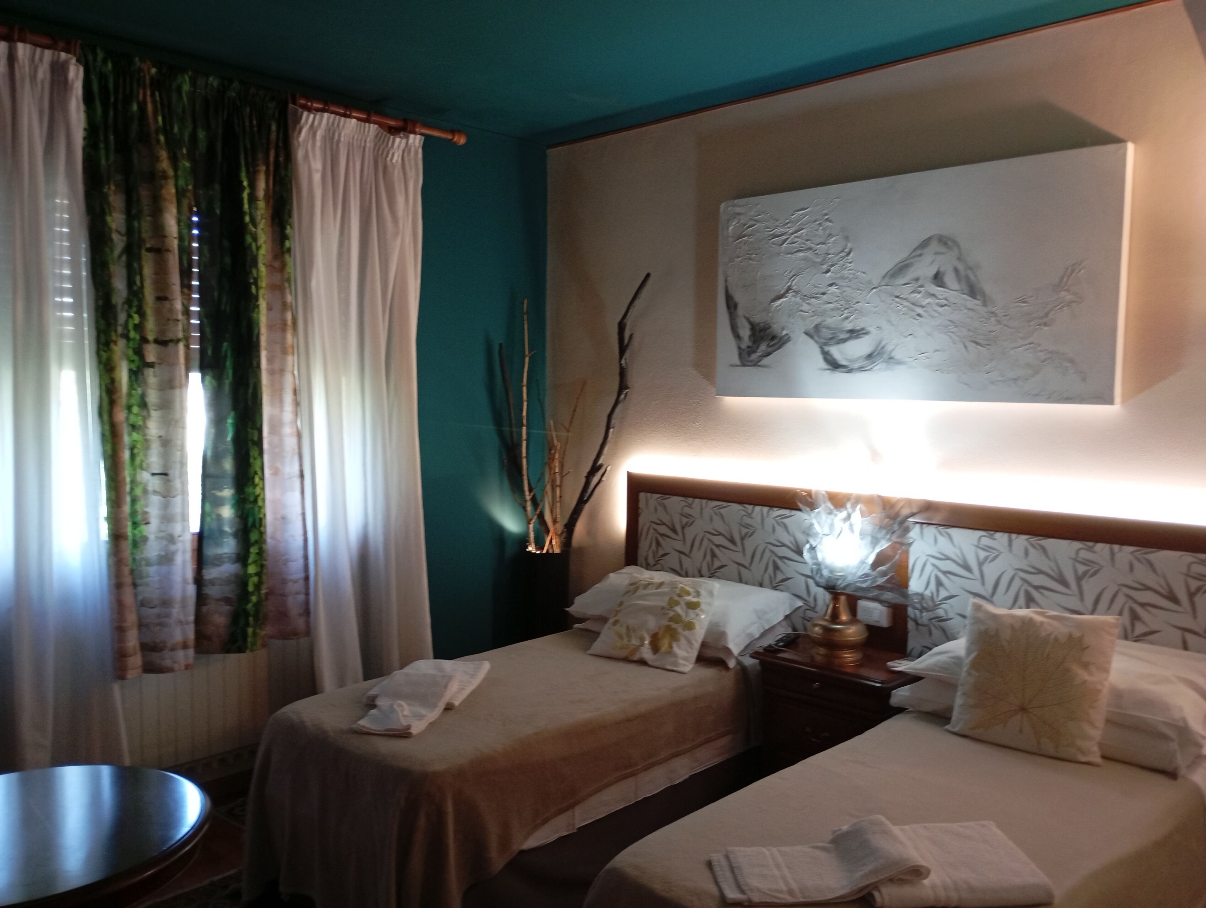 Una camera d'albergo con due letti, un comodino con una lampada, un quadro appeso al muro e una finestra con tende.