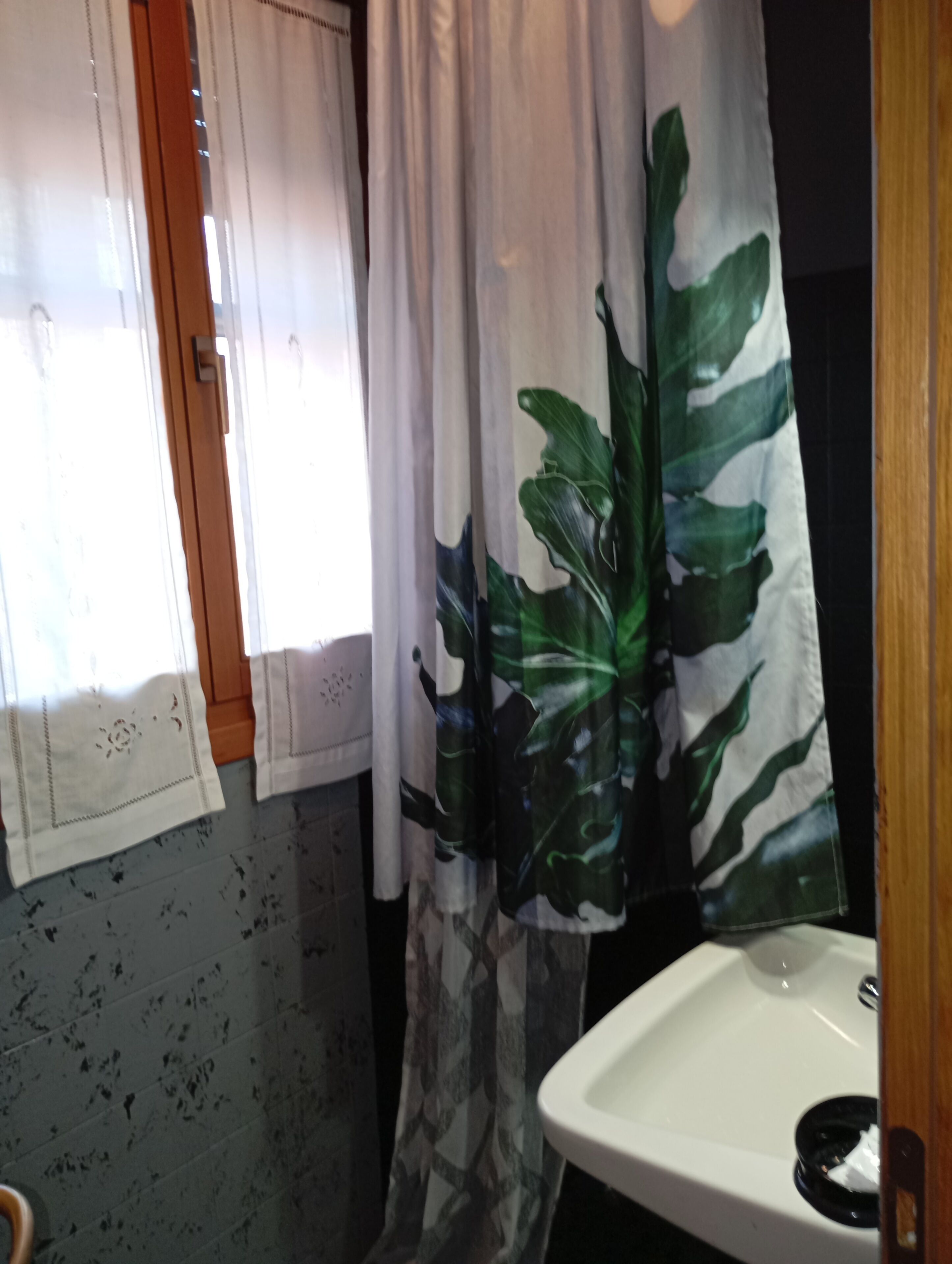 Une salle de bain avec un lavabo blanc, une fenêtre avec des rideaux blancs et un rideau de douche orné d’un motif de feuilles.
