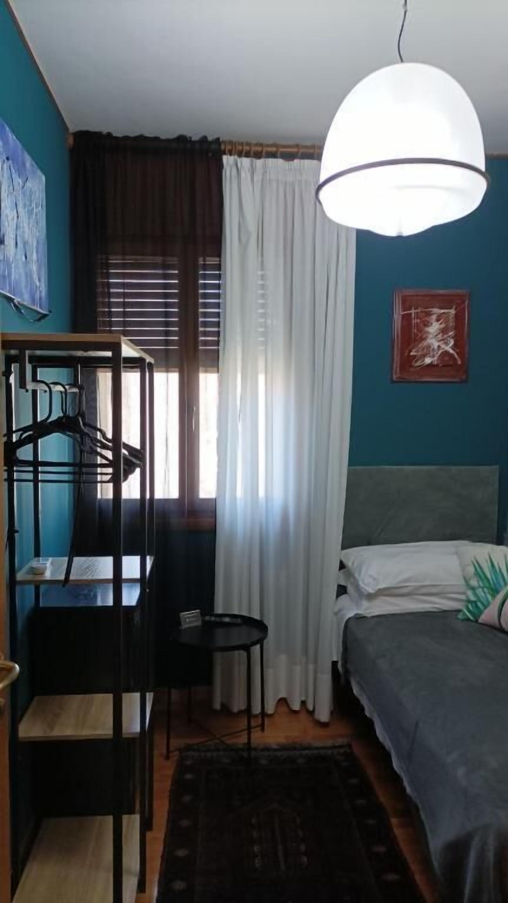 Une pièce comprenant un lit, un bureau, une chaise, un luminaire suspendu et une fenêtre avec des rideaux.