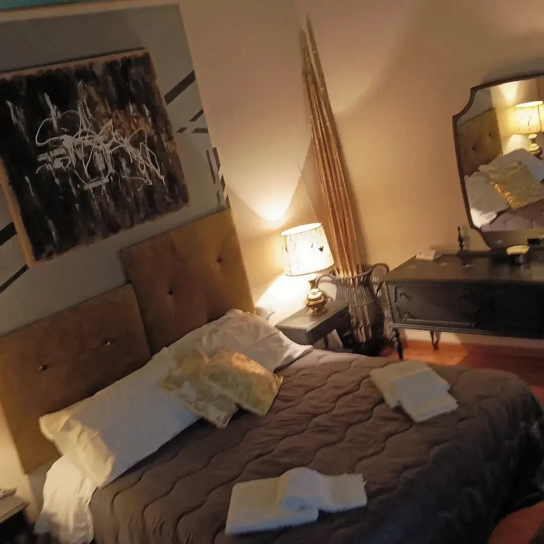 Quarto de hotel com uma cama, mesa de cabeceira, abajur, espelho e um quadro de arte abstrata na parede.