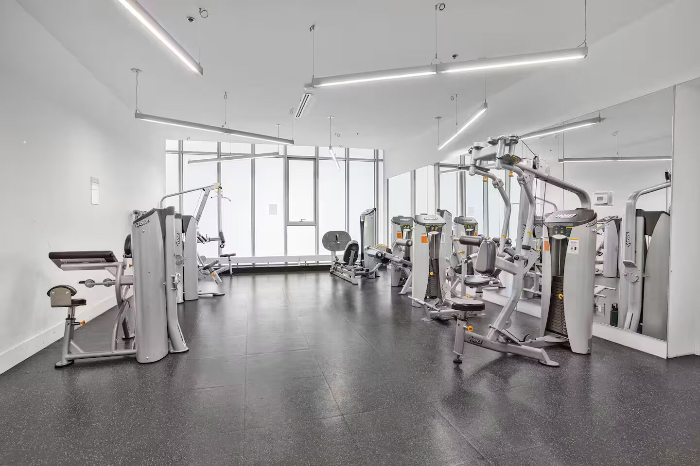 Une salle de sport moderne, dotée de divers appareils d’entraînement, de grandes fenêtres et d’un aménagement propre et spacieux.