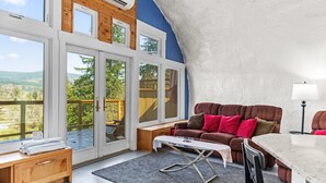 Una habitación con un gran ventanal corredero que da a una terraza, un sofá marrón con cojines rojos, una mesita y una lámpara.