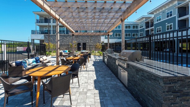 An outdoor patio with wooden tables and wicker chairs, a stone wall, and a grill area. - NEW Upscale Pool View Condo - Walk to Outlets + Near Beaches (Rehoboth Beach)