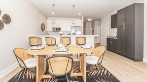 A modern kitchen with white cabinets, a central island, and a dining area with wicker chairs. - NEW Upscale Pool View Condo - Walk to Outlets + Near Beaches (Rehoboth Beach)