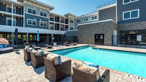 A modern building with a swimming pool, outdoor seating, and a covered area with umbrellas. - NEW Upscale Pool View Condo - Walk to Outlets + Near Beaches (Rehoboth Beach)