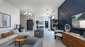 A modern living room with a sofa, armchairs, and a coffee table. There are two paintings on the walls, a chandelier, and a cabinet with decorative items. - NEW Upscale Pool View Condo - Walk to Outlets + Near Beaches (Rehoboth Beach)