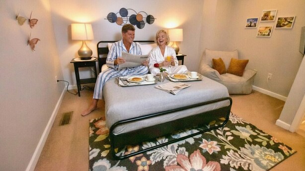 Un dormitorio con cama, mesitas de noche, lámpara, alfombra con motivos florales y adornos en la pared.