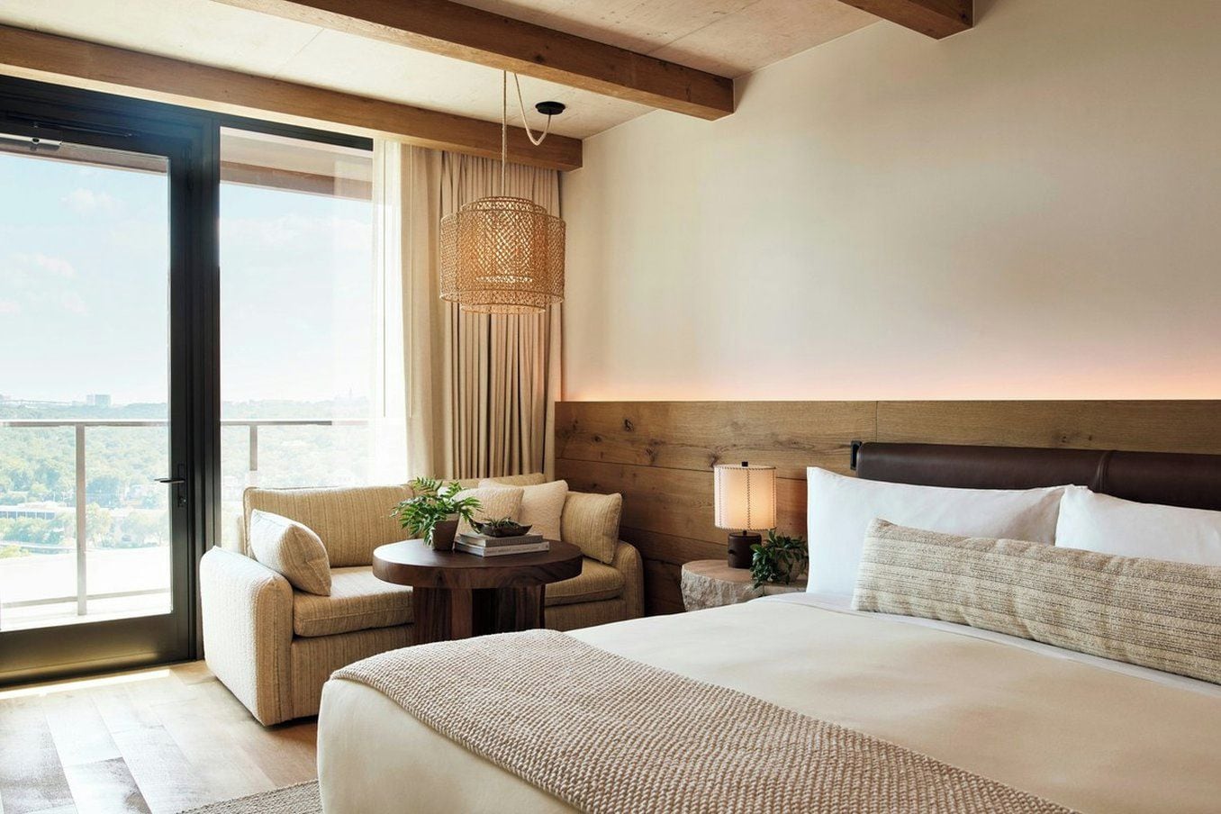 A hotel room with a bed, a sofa, a small table, a lamp, and a view through a large window.