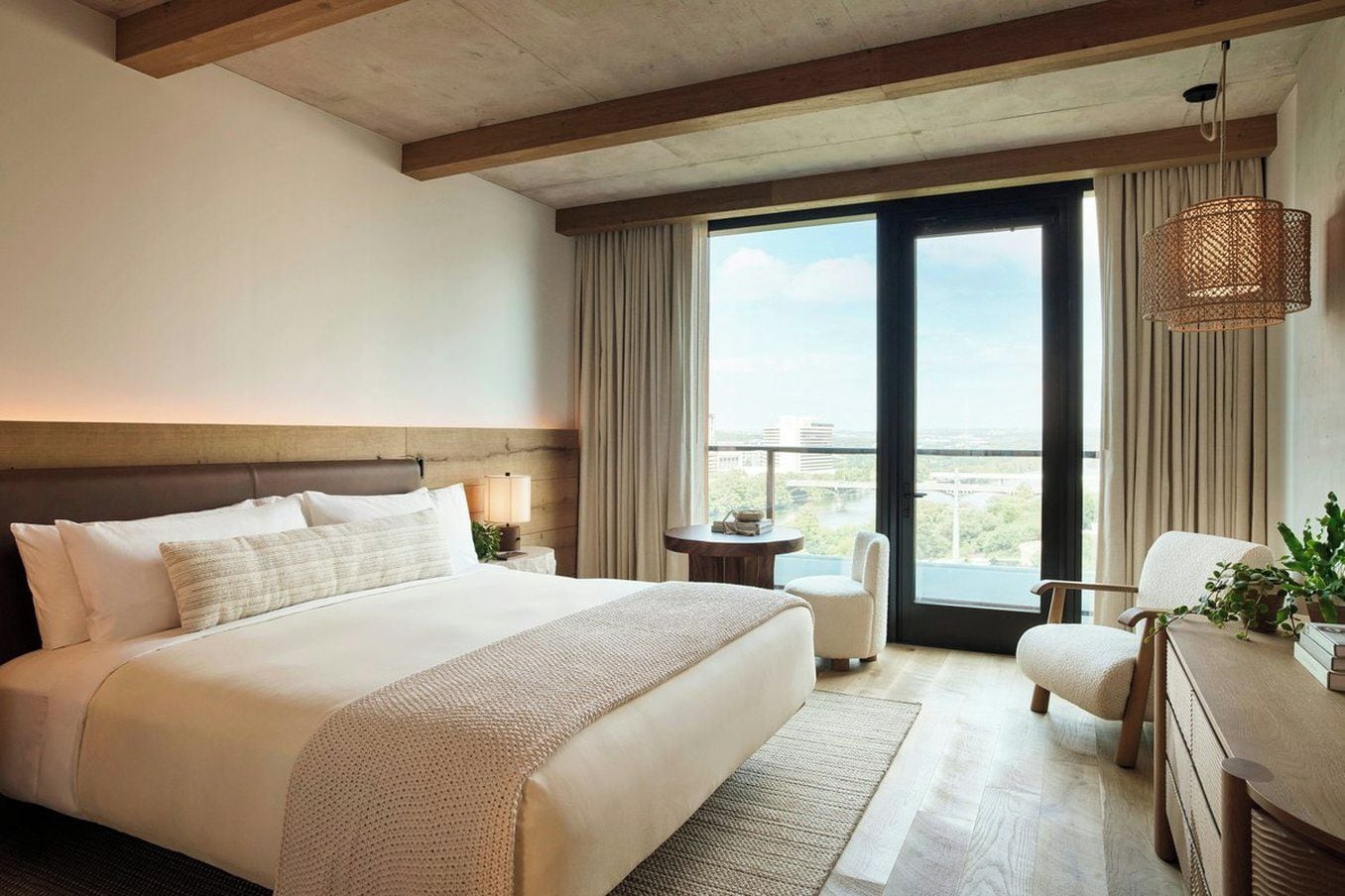 Una habitación de hotel con una cama grande, un área de descanso con una silla, una mesita y vistas de la ciudad a través de un ventanal amplio.