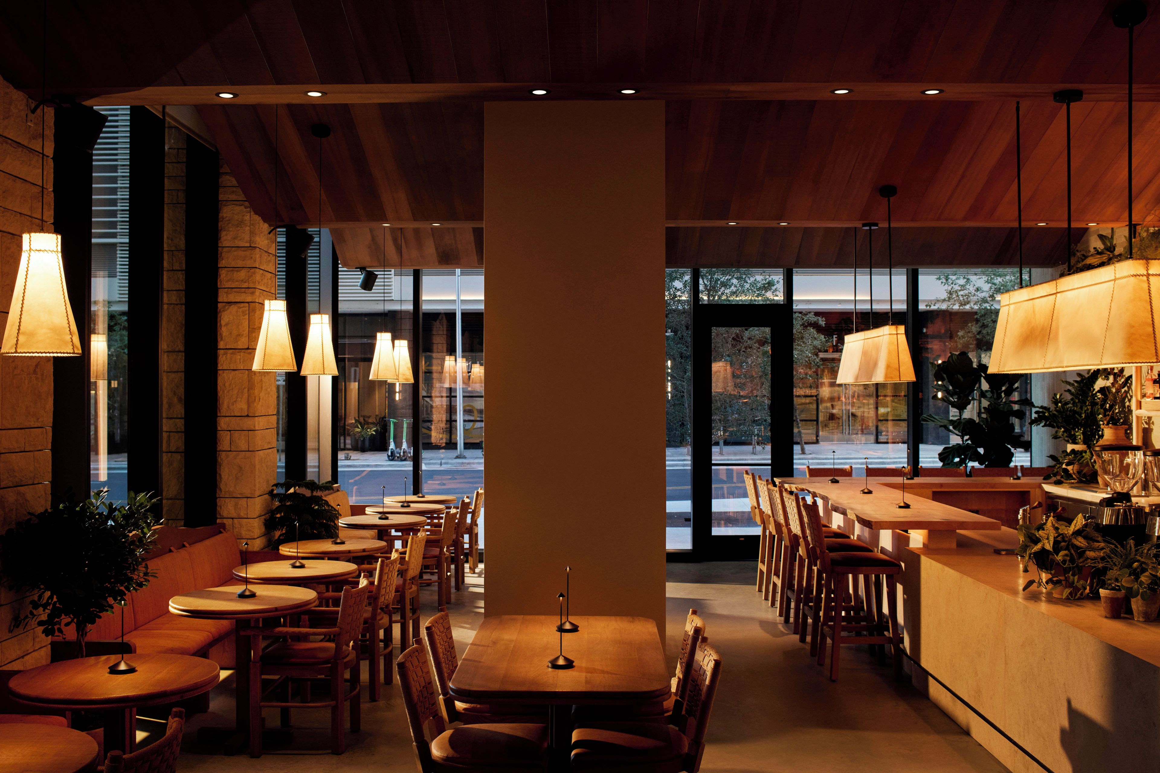 En restaurantindretning med træborde og stole, hængende lamper og udsigt gennem store vinduer.
