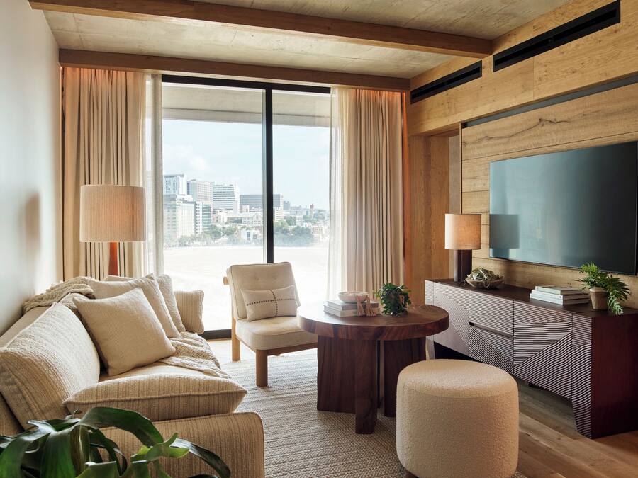 Une chambre d’hôtel moderne dotée d’une grande fenêtre, d’un canapé, d’un fauteuil, d’une petite table et d’un téléviseur fixé au mur.