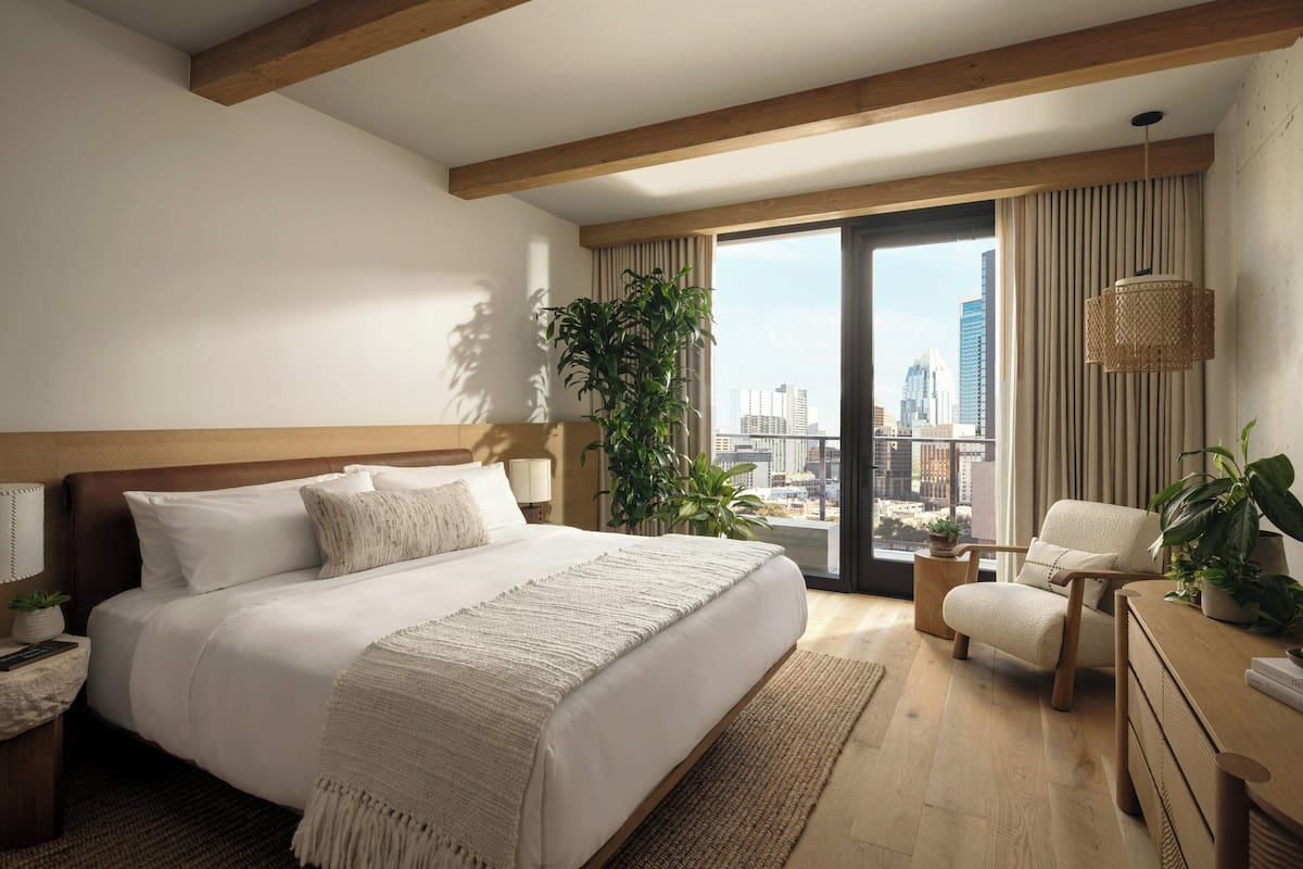 A modern hotel room with a large bed, a desk, a chair, and a view of the city skyline.