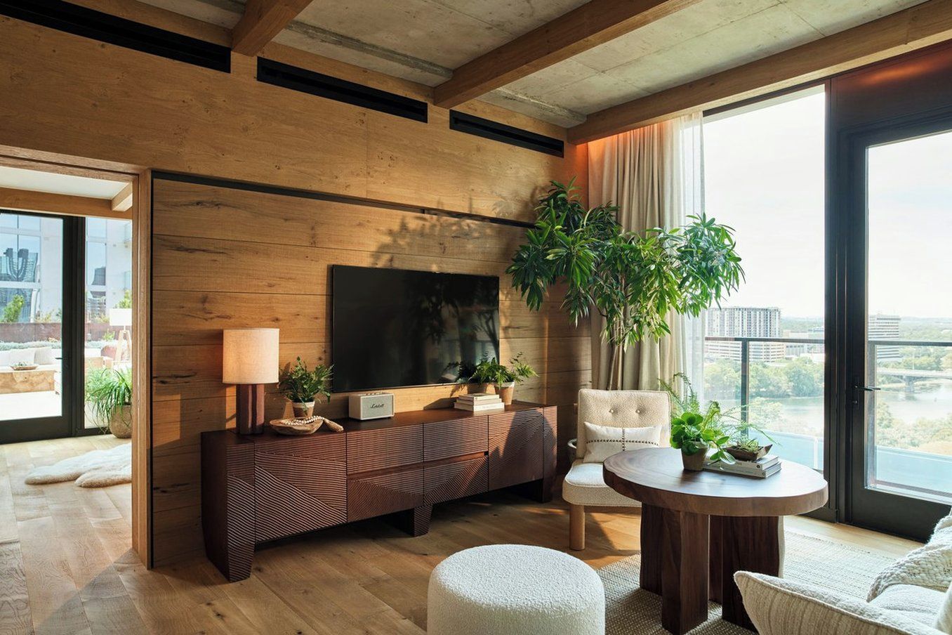 Un salón moderno con un televisor grande, paneles de madera, un sofá, una mesa de centro redonda y vistas de la ciudad.