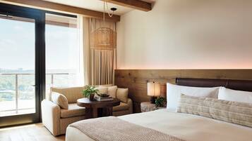 A hotel room with a large bed, a sitting area with a chair and a small table, a lamp, and a view of the city through the window.