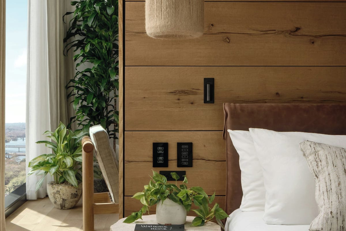 A bedroom with a bed, a bedside table with a welcome sign, a hanging light fixture, and a large potted plant.