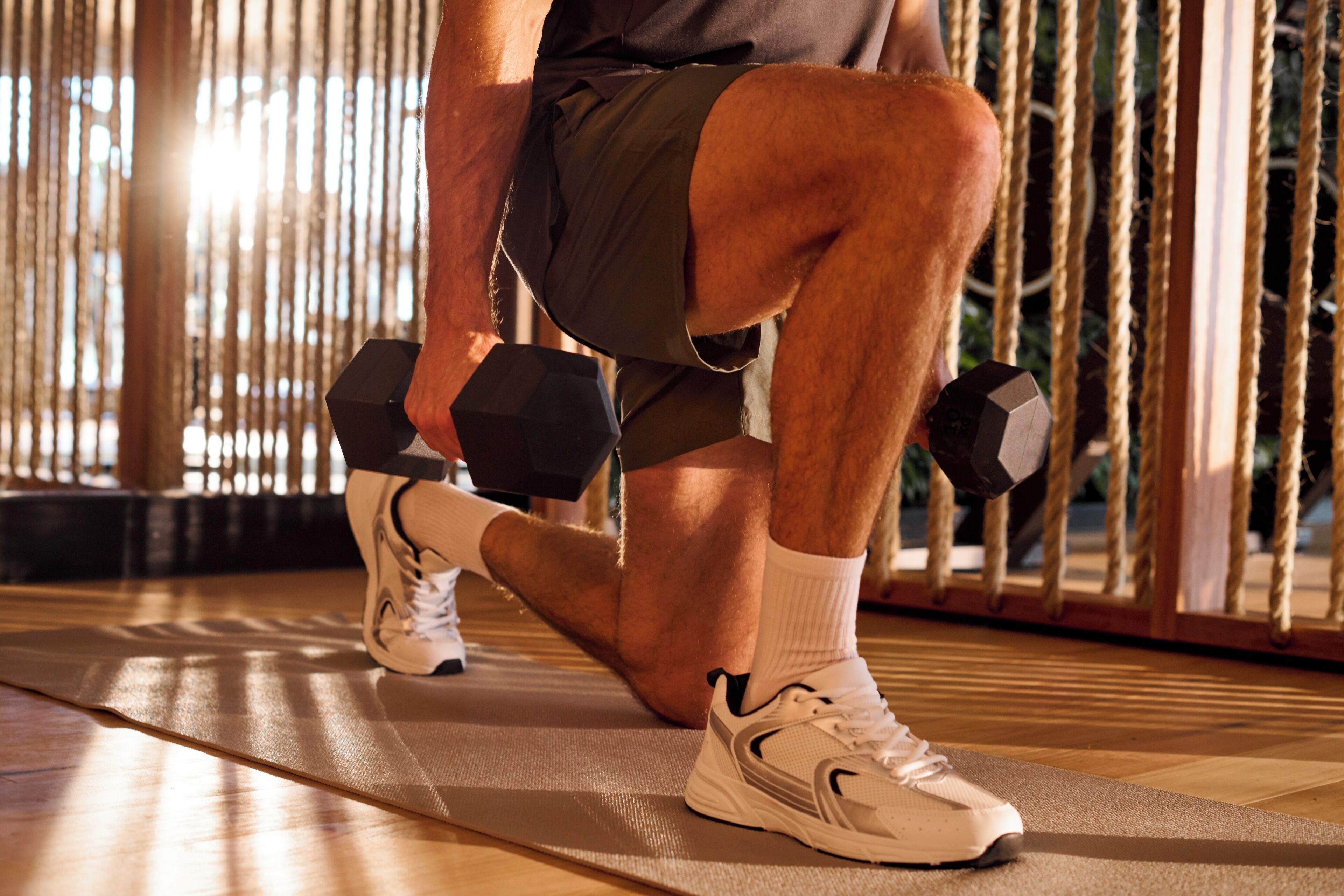 A person exercising with dumbbells on a mat in a room with a wooden floor and a rope barrier.