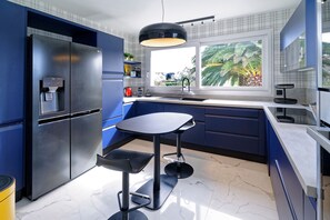 A modern kitchen with a built-in refrigerator, a round dining table, and a window with a view of palm trees. - Villa Goazy - 950m de la Plage (Plougonvelin)