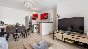 A modern living room with a flat-screen TV, dining area with chairs, a kitchen with red cabinets, and a washing machine. - La Brise d'Agde - 3km de la Plage (Agde)