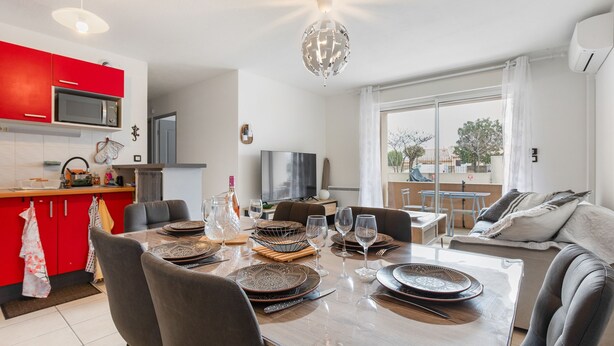 A modern kitchen with red cabinets, a dining table set with plates, glasses, and cutlery, and a living area with a sofa and a TV. - La Brise d'Agde - 3km de la Plage (Agde)
