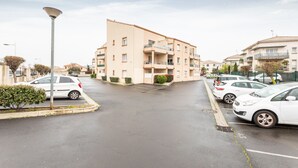 A parking lot with several cars, a lamp post, and a multi-story building with balconies. - La Brise d'Agde - 3km de la Plage (Agde)