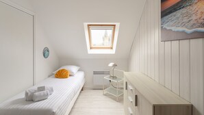 Quarto com claraboia, cama com roupa de cama branca, mesa de cabeceira com abajur e pintura de uma praia.