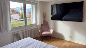 Ein Schlafzimmer mit einem Bett, einem Sessel, einem Fernseher, einem Fenster mit Vorhängen und einer Heizung.