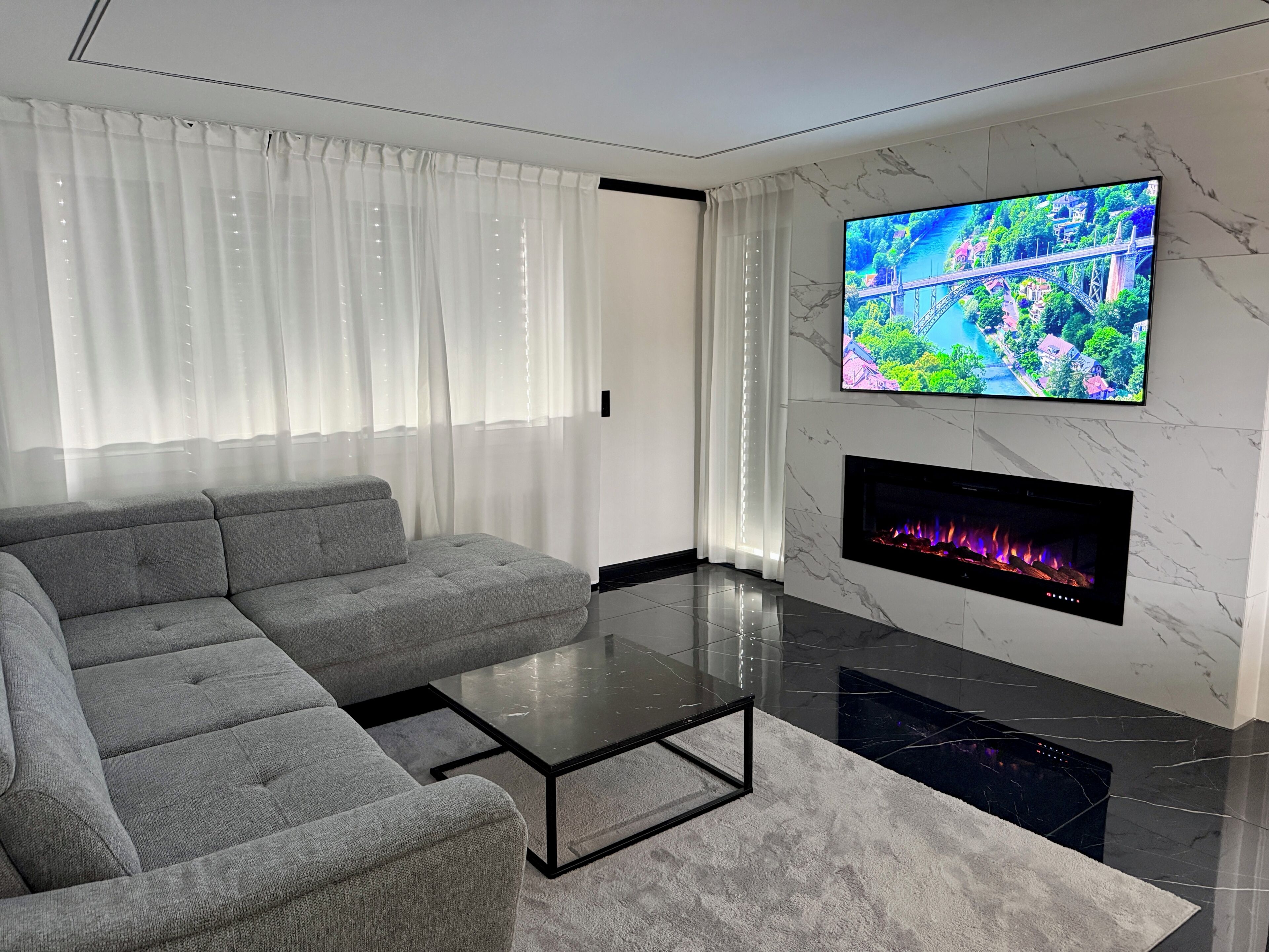 Een moderne woonkamer met een grijze modulaire bank, een glazen salontafel, een open haard en een televisie bevestigd aan een marmeren wand.