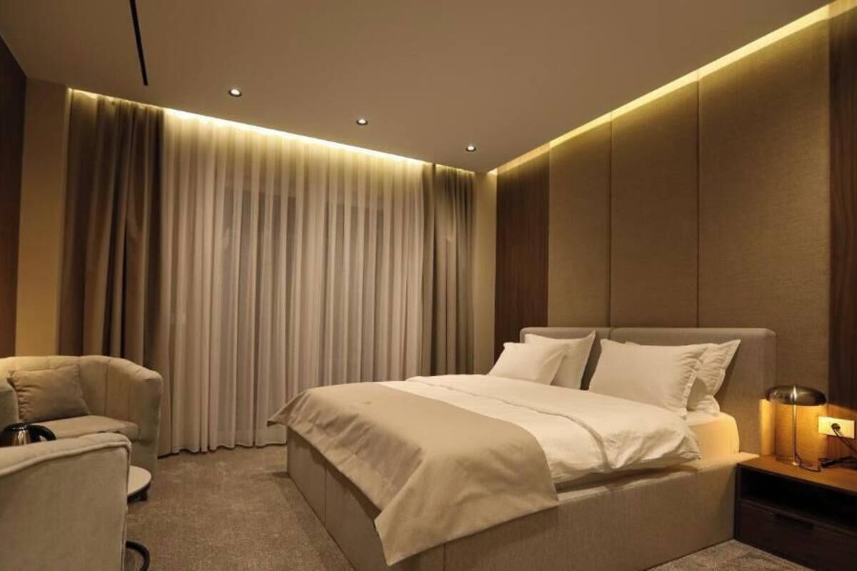 A modern hotel room with a large bed, a bedside table with a lamp, a chair, and a large window with curtains.