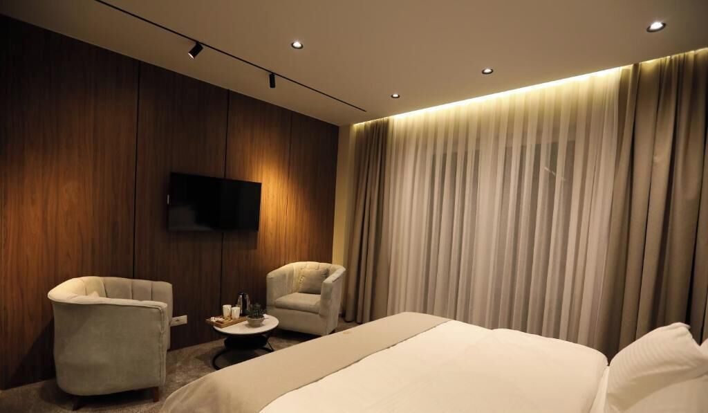 Una camera d'albergo con un letto, due poltrone, un tavolino, una TV a schermo piatto fissata al muro e un'ampia finestra con tende.