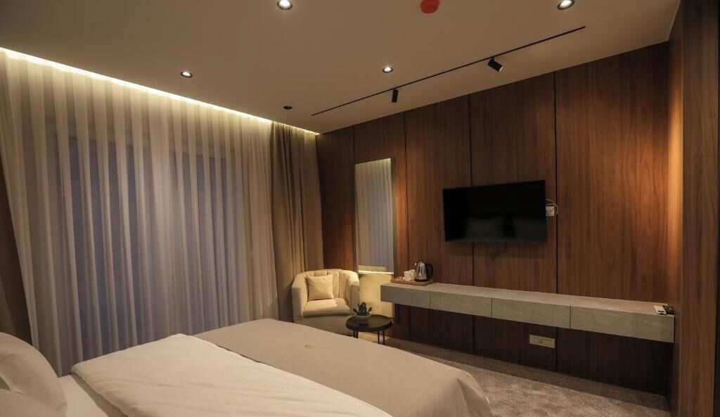 Habitación de hotel con cama, zona de estar con una silla, una mesita y un televisor montado en la pared, además de detalles decorativos en paneles de madera.