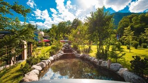 A pond with a stone border, surrounded by trees and a building with a red umbrella. - Resort Anës Drinit  (Radac)