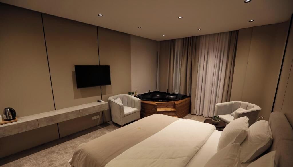 Una habitación de hotel moderna con una cama grande, dos sillones, un televisor de pantalla plana, un jacuzzi integrado y una mesita de madera.
