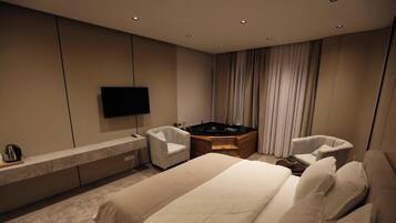 A modern hotel room with a large bed, two armchairs, a flat-screen TV, a built-in hot tub, and a wooden side table.