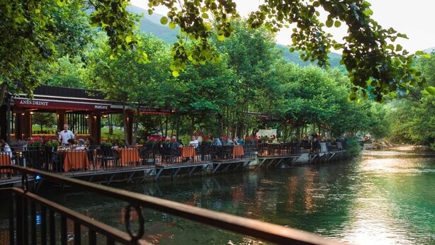 A riverside restaurant with outdoor seating, a wooden deck, and a view of the water. - Resort Anës Drinit  (Radac)