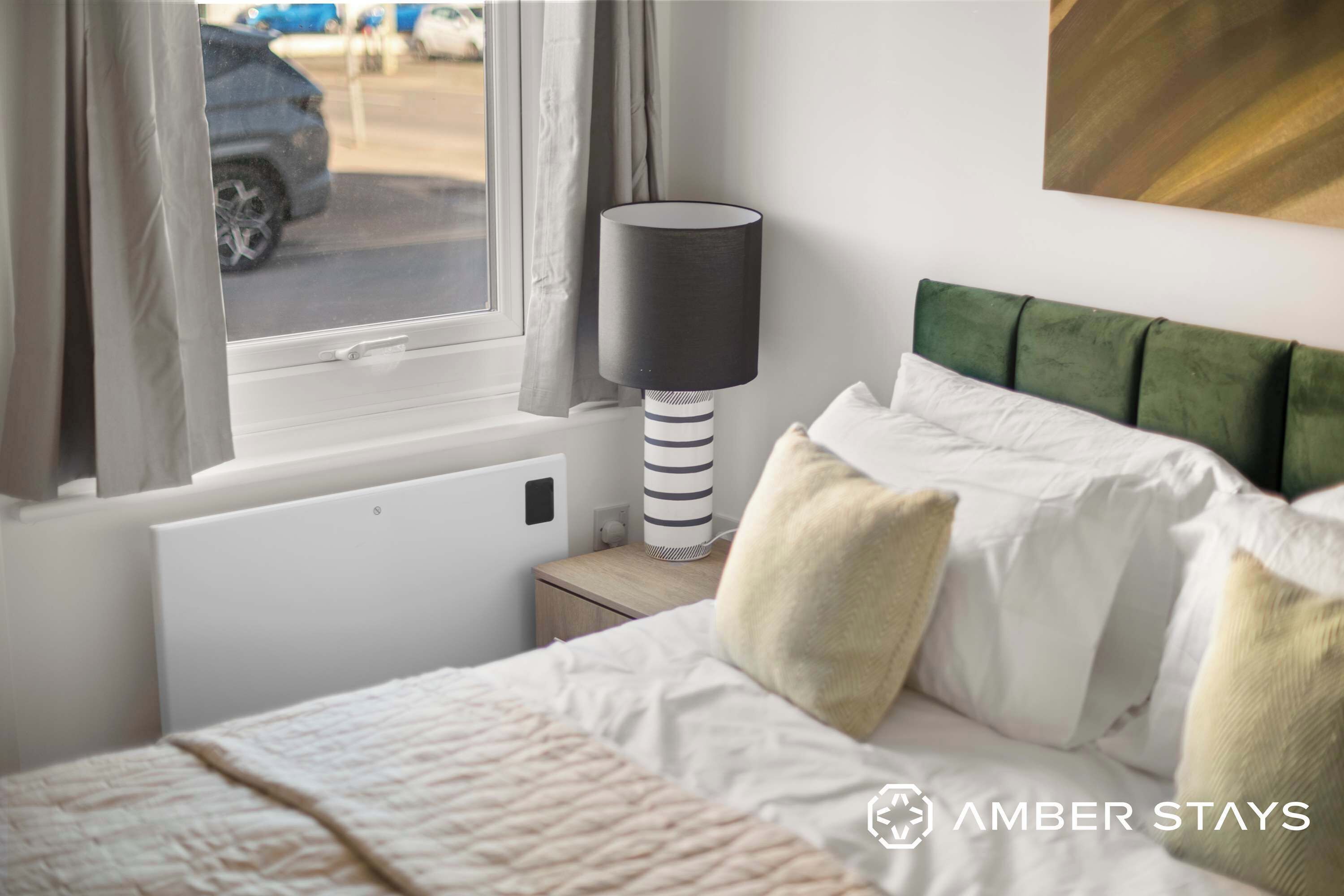 A cozy bedroom featuring a plush green velvet headboard, crisp white linens, and a modern striped lamp set against a bright window.