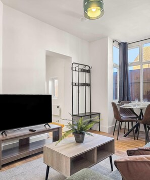 A modern living room with a flat-screen TV, a coffee table with a plant, a dining area with chairs, and a large window with curtains. - 234 Birstol Rd Apt, Gloucester (Gloucestershire)