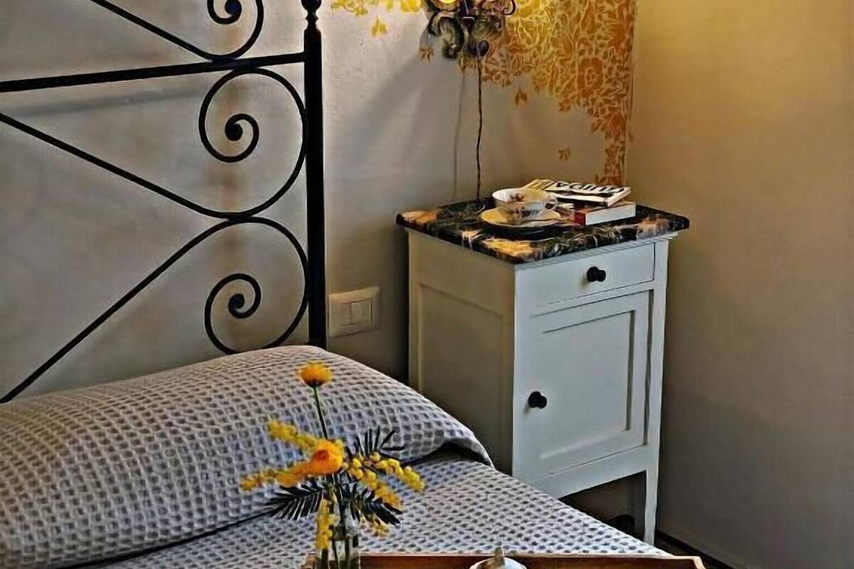 Un lit bien fait, avec un mur décoré d’un motif floral, une table de chevet sur laquelle se trouve un plateau avec du thé et des accessoires, et une lampe fixée au mur.