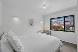 Ein Schlafzimmer mit Bett, Schreibtisch, Stuhl, Fenster mit Bergblick und einem gerahmten Bild an der Wand.