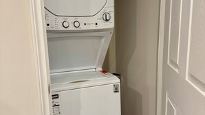A compact, white, stacked washer and dryer unit in a narrow hallway with a wooden floor and a white door. - Newly constructed apartment located near Canada’s Wonderland, Vaughan Mills Mall (Vaughan)