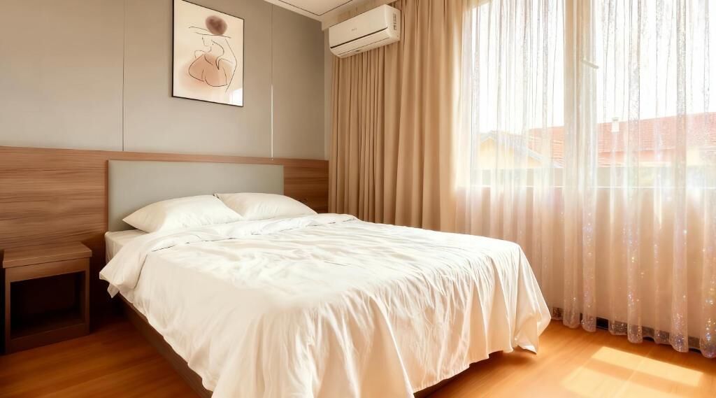 A neatly made bed in a hotel room with a headboard, an air conditioning unit, and sheer curtains.