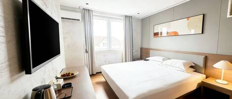 A modern hotel room with a flat-screen TV, a bed with white linens, a bedside table with a lamp, and a view of the outside through a window.