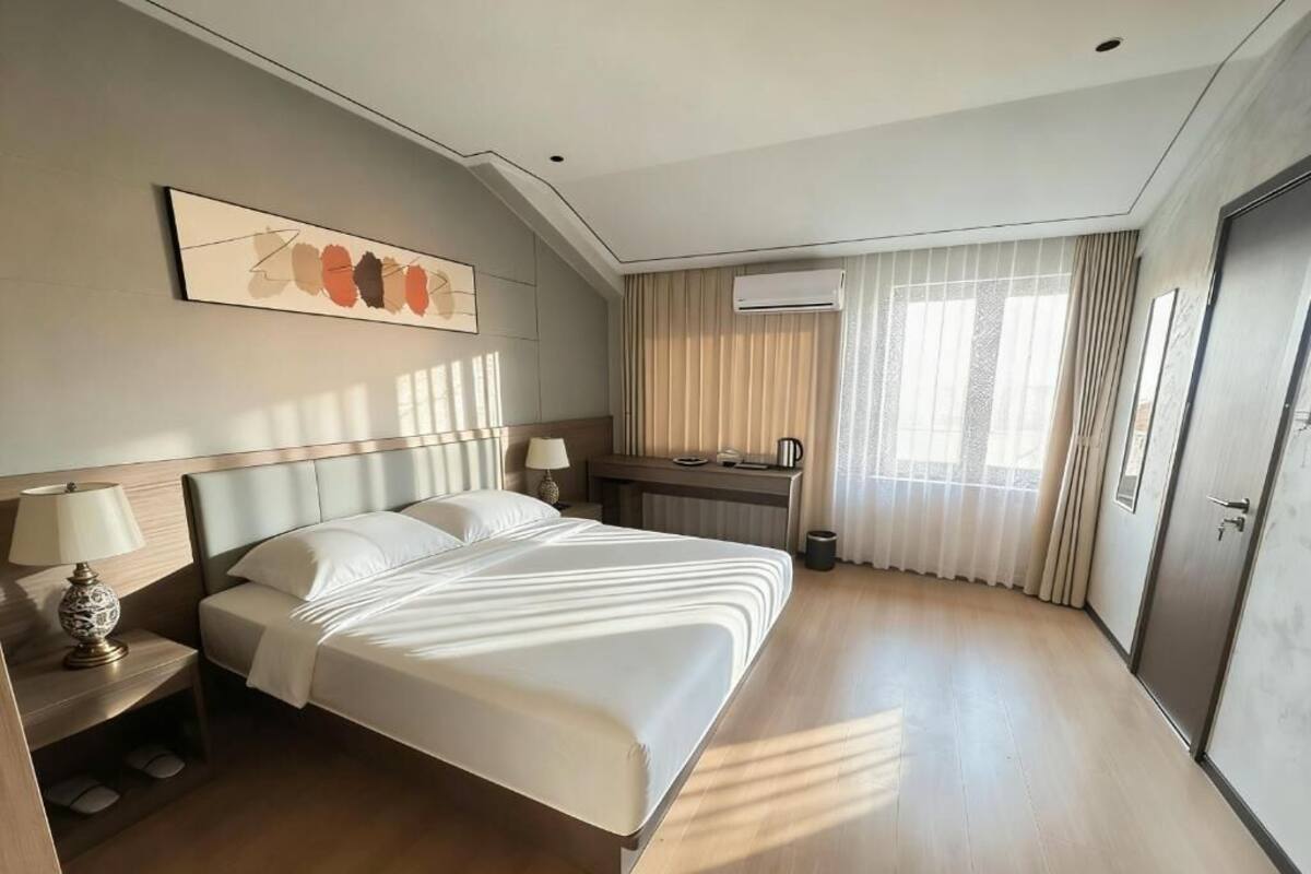 Habitació d'hotel amb un llit gran, una taula de treball, una cadira, una làmpada, un aparell d'aire condicionat i una finestra amb cortines.