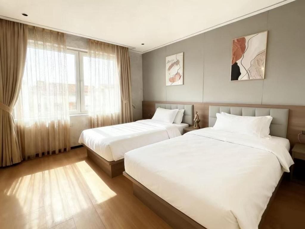 A hotel room with two beds, a large window with curtains, and artwork on the walls.
