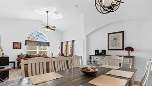 Ein Essbereich mit einem langen Holztisch, weißen Stühlen, einem Deckenventilator und einem großen Fenster mit Jalousien.